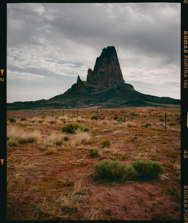 A large rock formation rises from a desert landscape under a cloudy sky.
