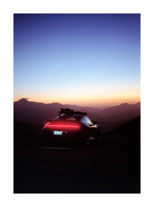 A car with a roof rack and glowing red taillights is parked on a mountain road at dusk.