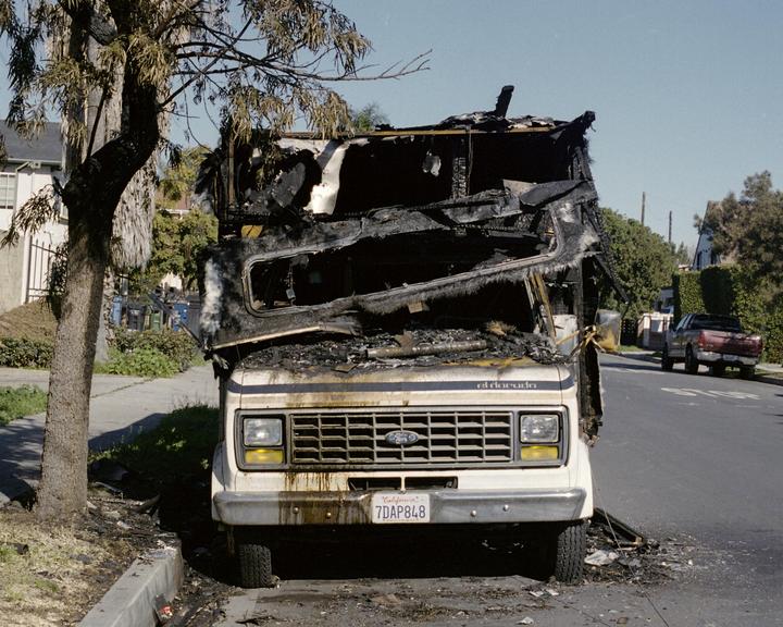 A burnt camper van is parked on the side of a street next to a tree.