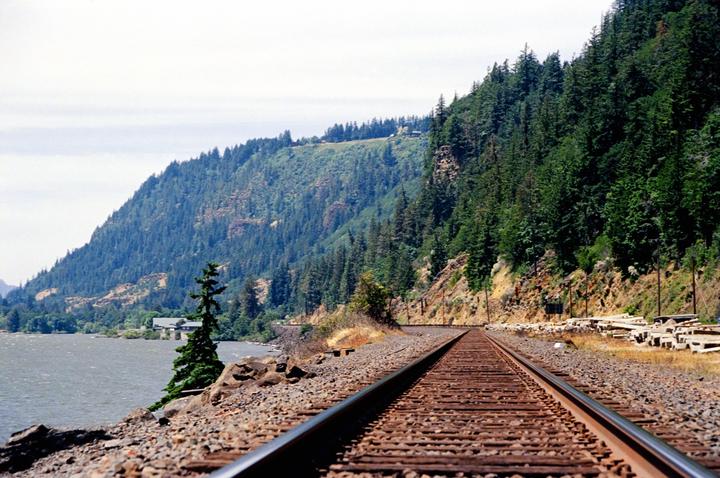 A train track runs along a river next to a forested mountain.