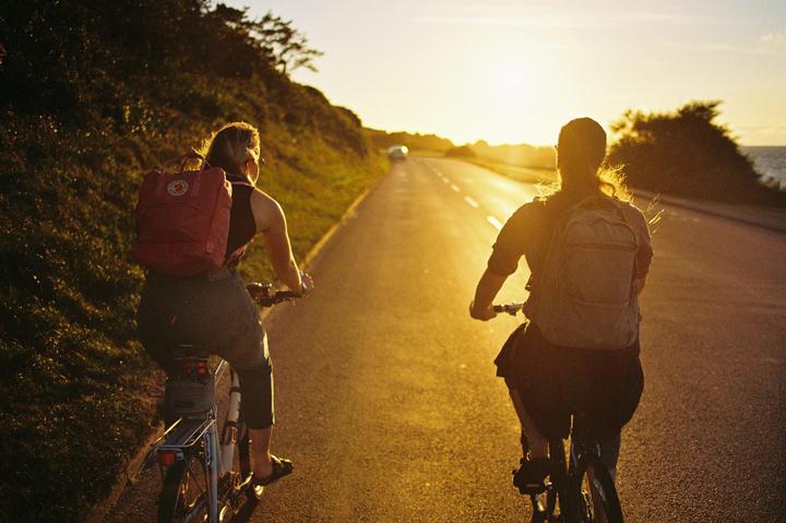 Two women ride bicycles down a road at sunset, with the ocean visible in the distance.
