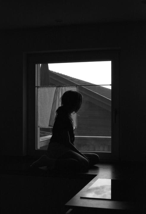 A girl sits silhouetted in front of a window, looking out at a building with a sloped roof.