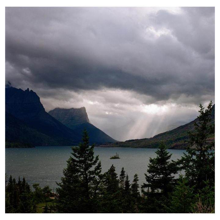 A lake with a small island is surrounded by mountains under a cloudy sky with sun rays breaking through.