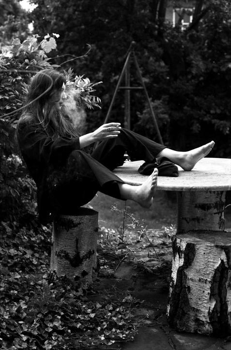 A woman with long hair sits barefoot on a tree stump, smoking a cigarette in a garden.