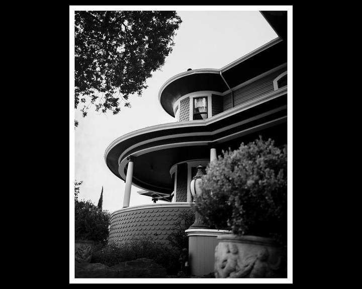 A black and white photograph shows a Victorian house with a rounded porch and a tree in the upper left corner.