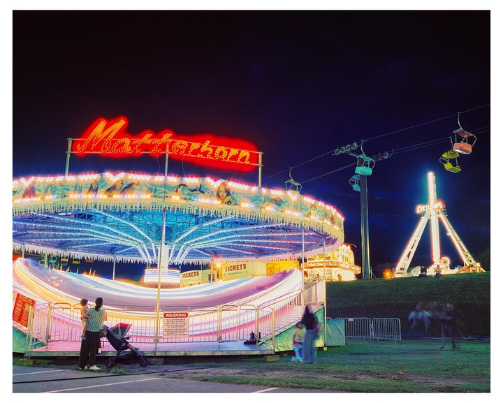 A fairground at night features a spinning Matterhorn ride with blurred lights, a ski lift, and a Cyclops ride in the background, with people watching the rides.