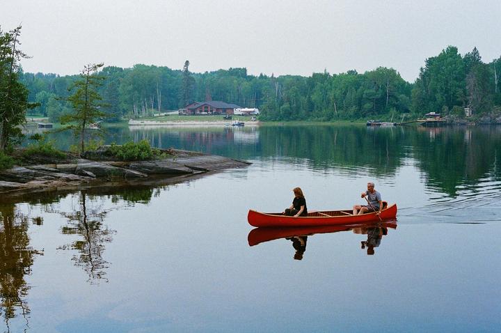 A couple paddles a red canoe on a calm lake surrounded by a forest and a distant building.