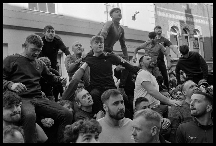 A large group of men are gathered in a street, with some men sitting on the shoulders of others.