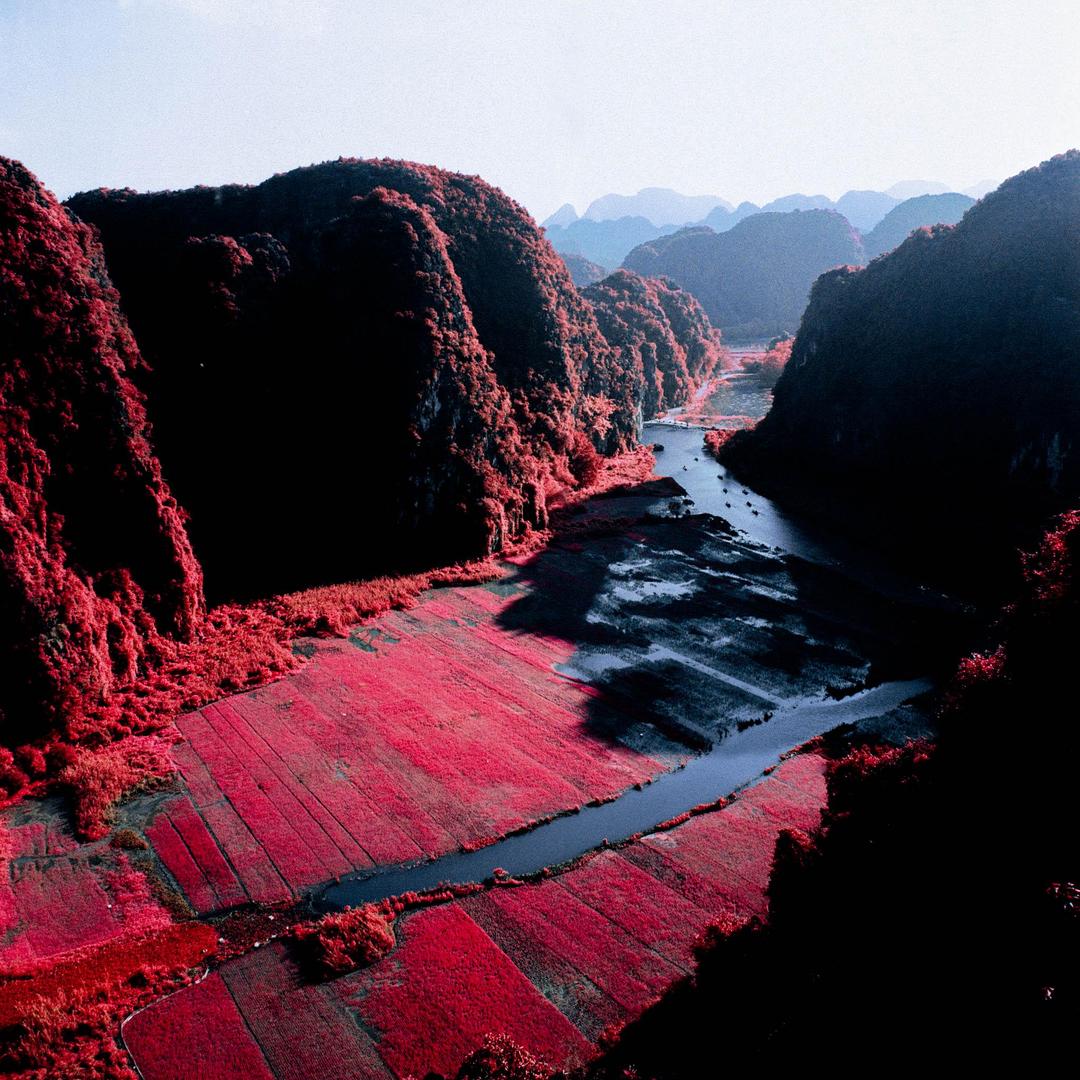 A river winds through a valley with red foliage and mountains under a bright sky.