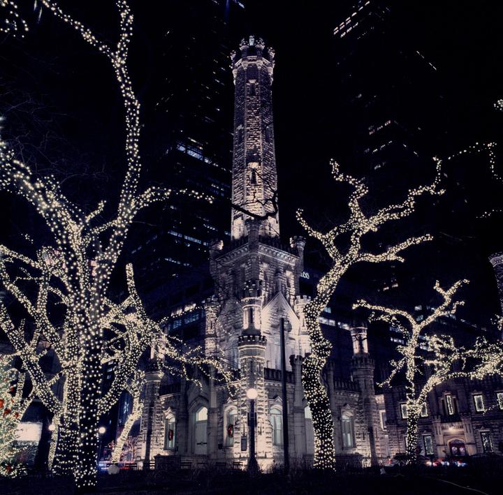 A tall stone tower is illuminated at night, surrounded by trees wrapped in string lights.