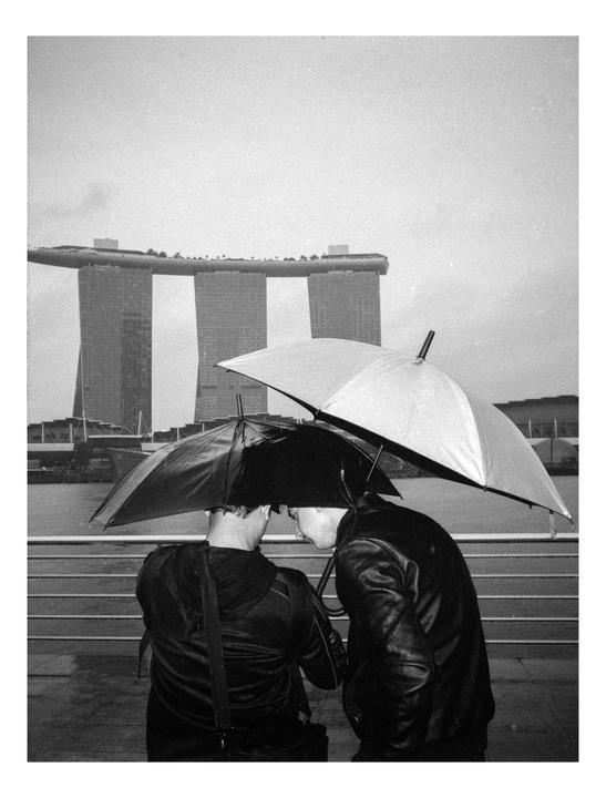 Two men stand under umbrellas with a large building in the background.