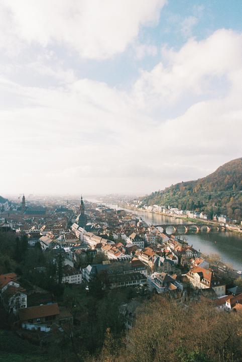 A panoramic view shows the city of Heidelberg with its buildings and a river flowing through it, surrounded by hills.