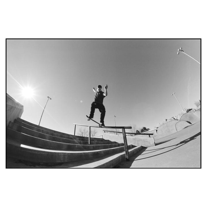 A man on a skateboard grinds a rail at a skatepark under a bright sun.