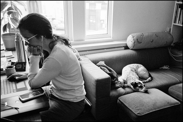 A woman works on a laptop at a desk while a dog sleeps on a sofa next to her.