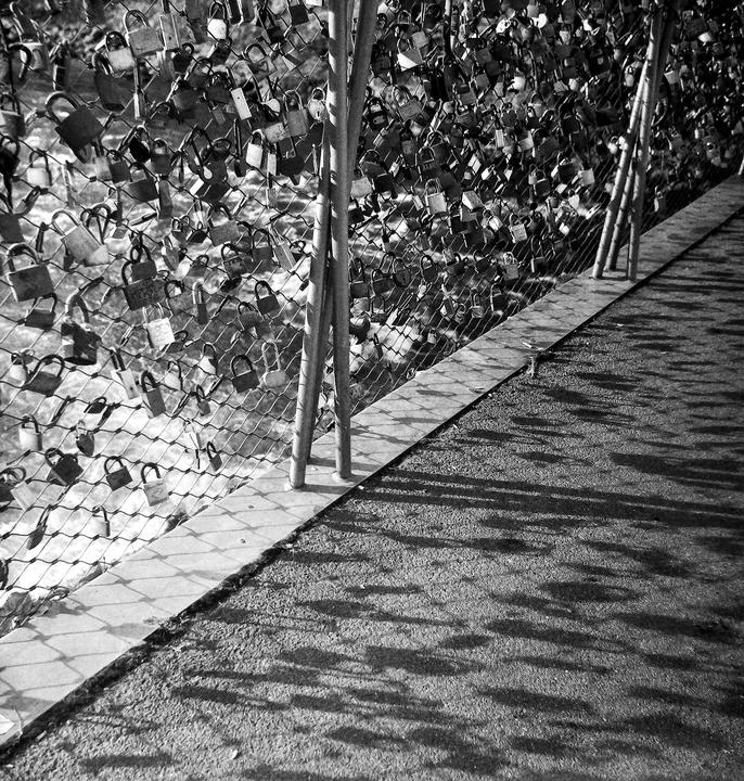 A fence covered in padlocks stands next to a path with shadows cast upon it.