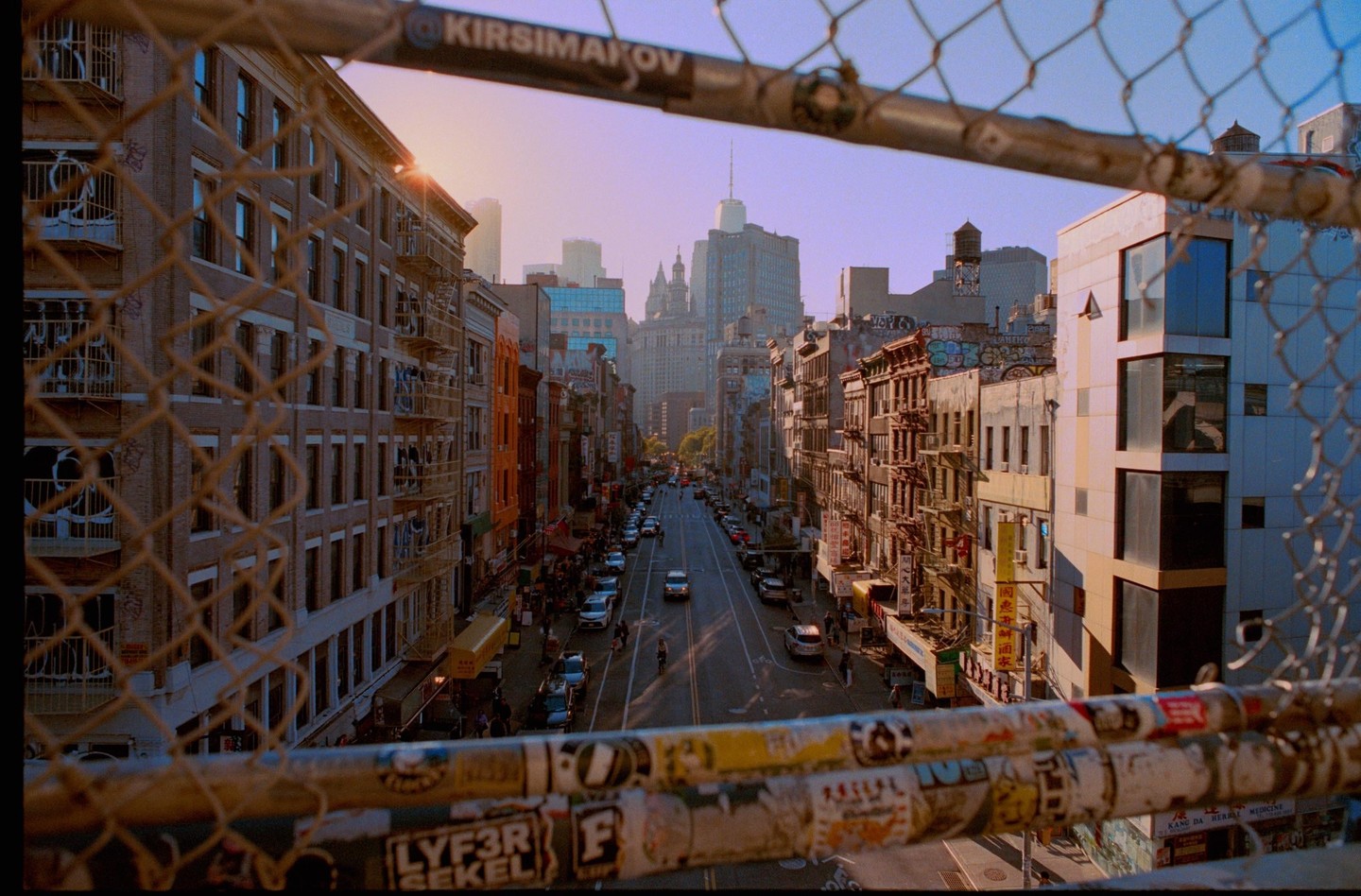 A street in New York City is viewed through a chain link fence with buildings and cars lining the road.
