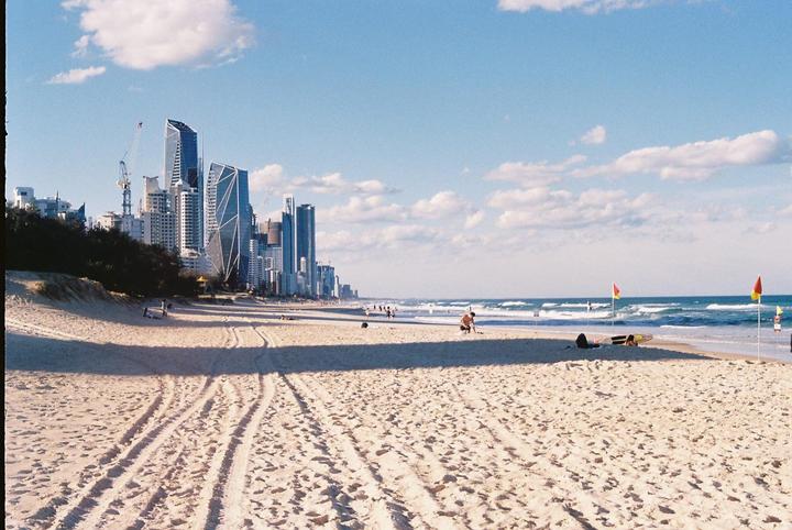 A wide sandy beach with tire tracks leads to the ocean, with a city skyline in the distance under a blue sky with clouds.