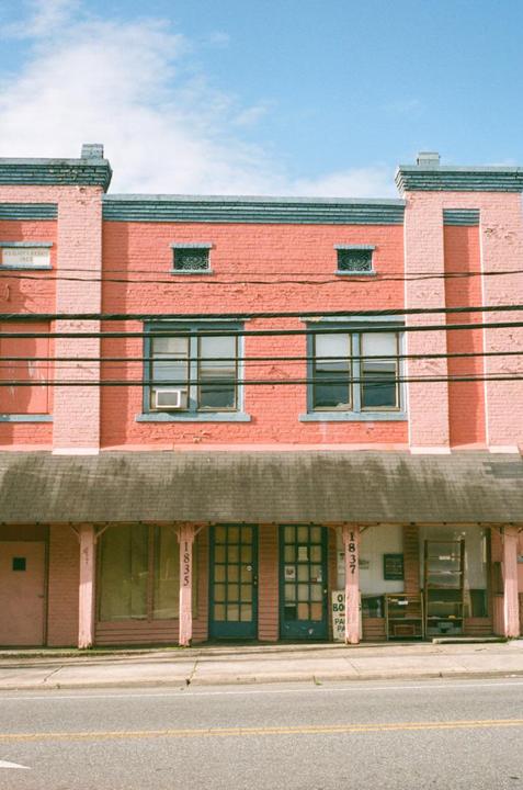 A two-story brick building with a faded pink facade and blue trim stands beside a road under a clear sky.
