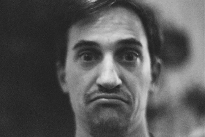 A man with a mustache and goatee looks directly at the camera with a sad expression.