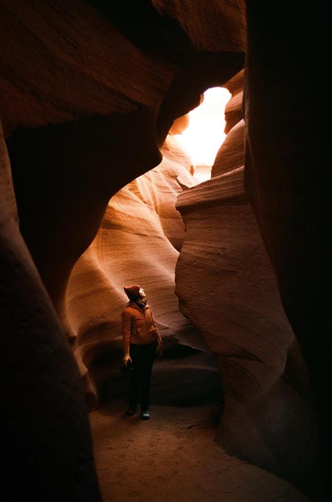 A woman in a red hat and orange jacket looks up at the light coming into a slot canyon.