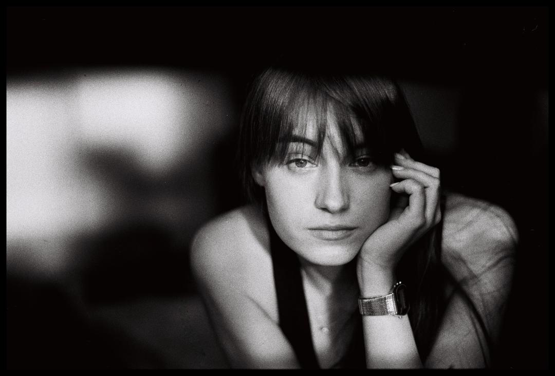 A woman with bangs and a watch rests her chin on her hand while looking at the viewer.