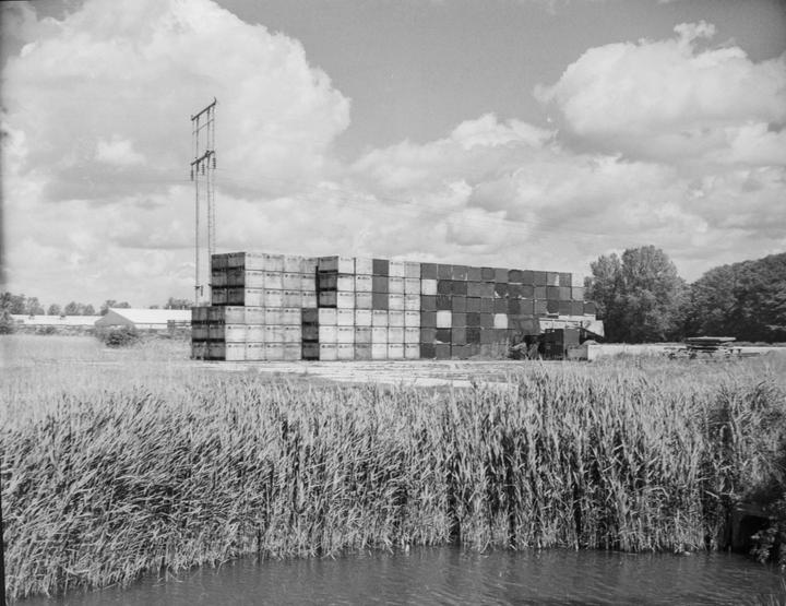 A large stack of boxes sits next to a body of water with tall grass in the foreground and a power line in the background.