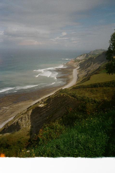 A long, narrow beach with waves breaking on the shore is visible from a high cliff covered in green grass and trees.