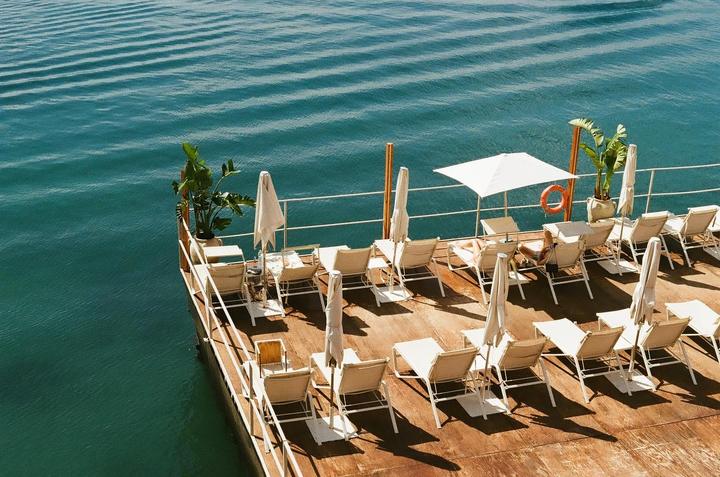 A wooden deck with lounge chairs and umbrellas extends over blue water.