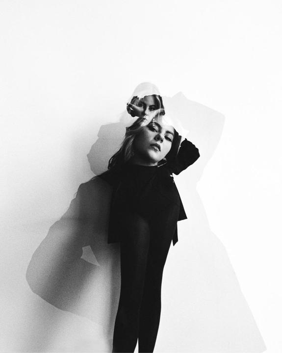 A woman with long hair is captured in a double exposure against a white wall, creating a ghostly effect.