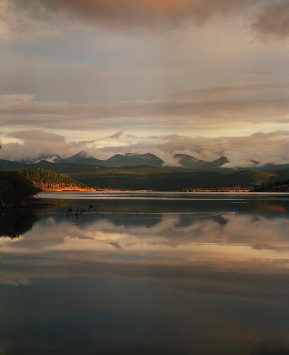 A lake reflects the cloudy sky and mountains with trees on the shore.