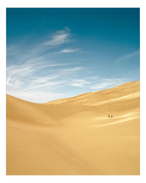 Two people stand in the distance on a vast sand dune under a blue sky with wispy clouds.