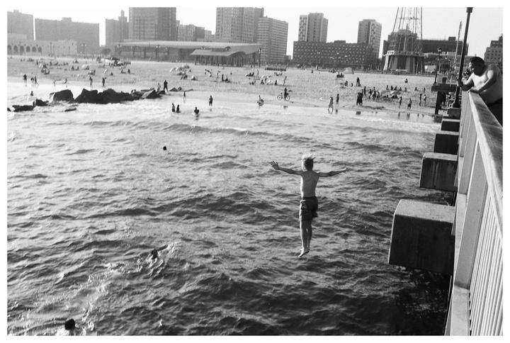 A boy jumps from a pier into the ocean while a man watches and people relax on a distant beach with buildings in the background.