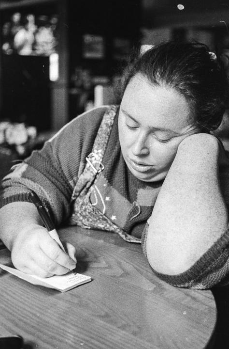 A woman with her eyes closed is leaning on her arm and writing on a small piece of paper at a wooden table.