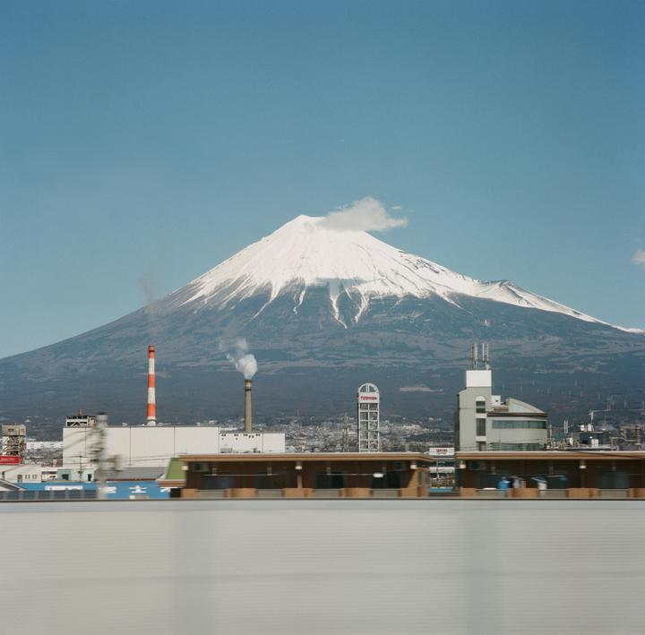 Mount Fuji with a snow-capped peak rises behind industrial buildings and a moving foreground.