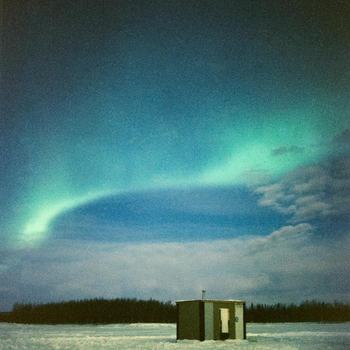 A small hut sits on a snowy field under a sky illuminated by the northern lights.