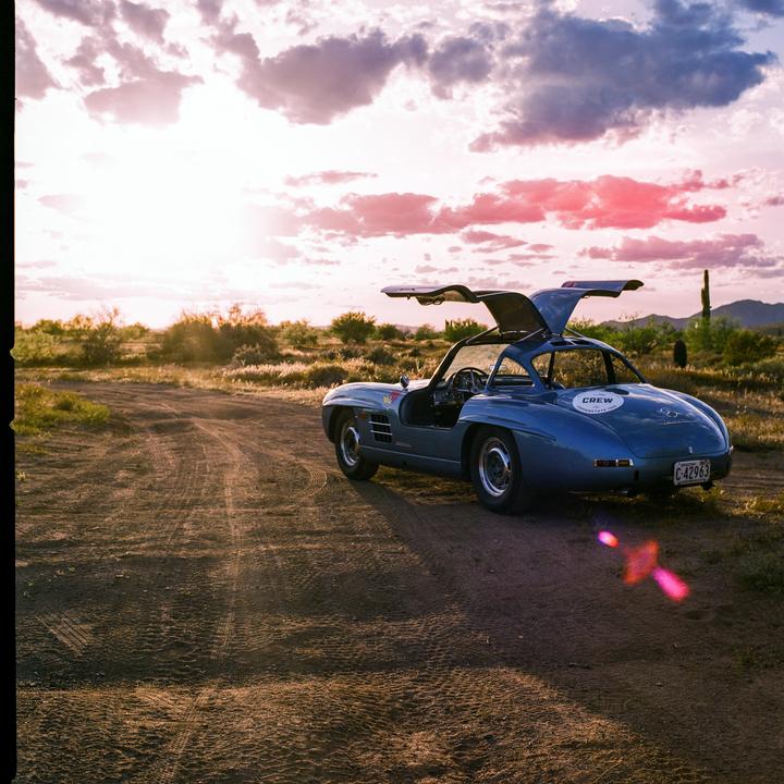 A blue car with its doors open is parked on a dirt road at sunset.