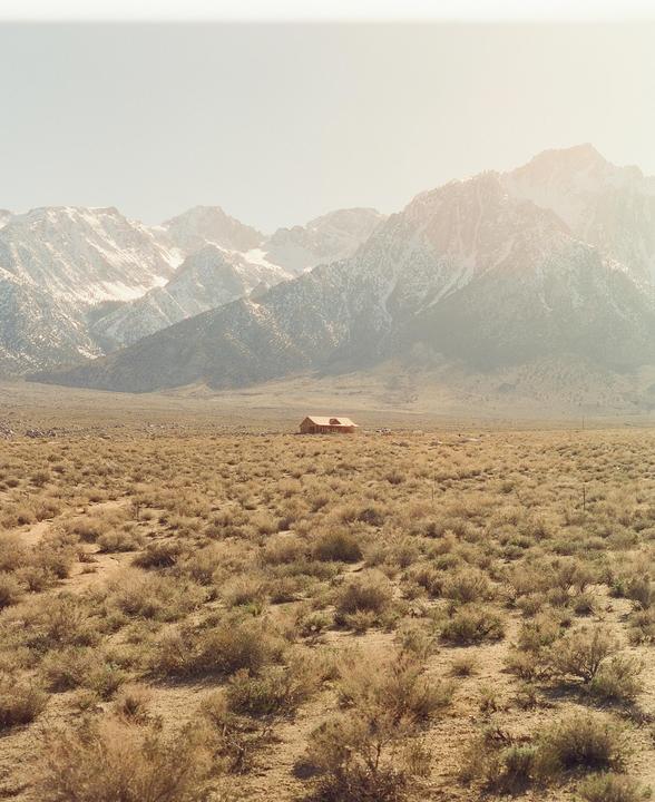 A wooden house sits in a vast, arid landscape with snow-capped mountains in the background.