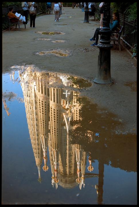 The Sagrada Familia is reflected in a large puddle on a dirt path where people are walking and sitting.