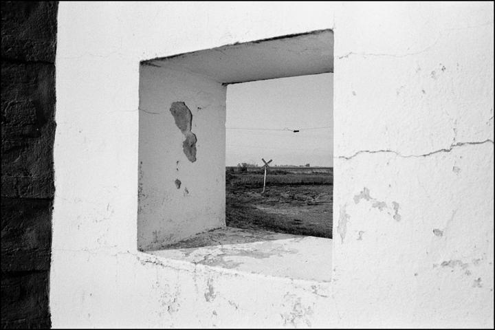 A railroad crossing sign stands in a field as seen through a window opening in a weathered building.