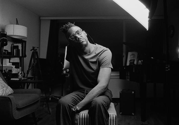 A man with glasses and dreadlocks sits on a striped stool in a room with a piano and a bright light.