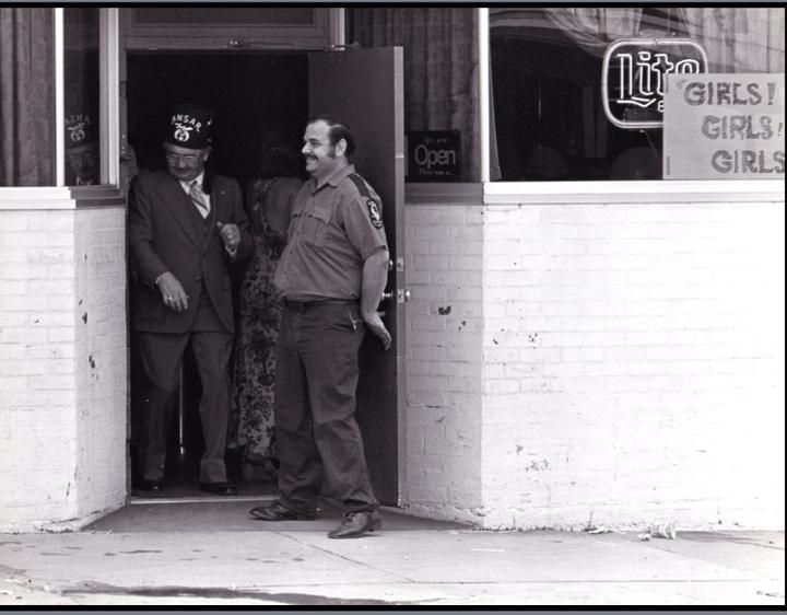 A man in a fez and suit exits a building next to another man in a uniform.