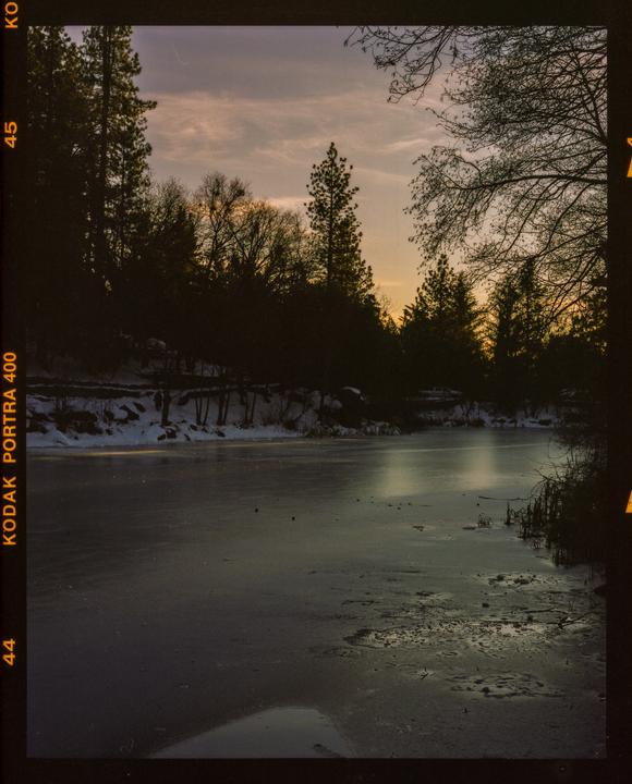 A frozen lake is surrounded by snow-covered banks and silhouetted trees under a colorful sky at dusk.