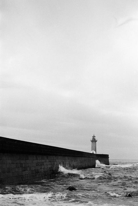 A lighthouse stands at the end of a stone pier with waves crashing against it.