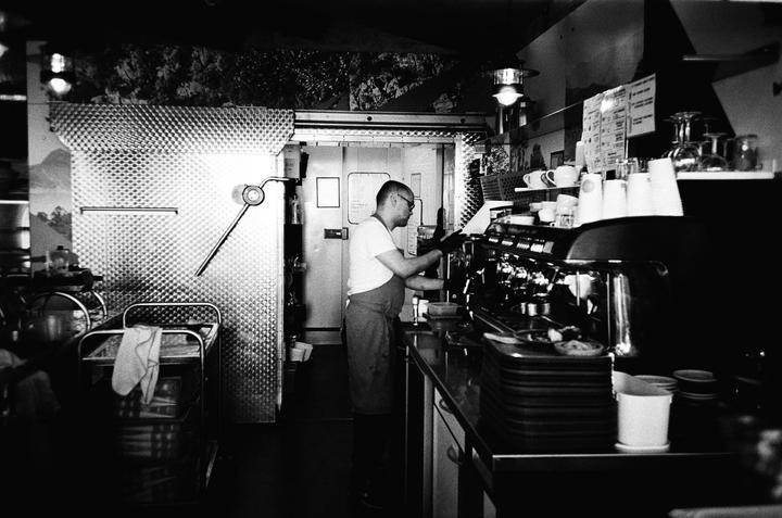 A man wearing an apron and glasses stands behind a counter with a coffee machine and stacks of cups.