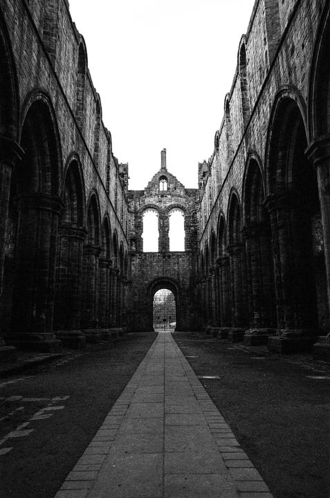 A long stone path leads through the ruins of an abbey toward an arched gate.