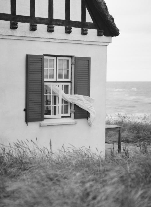 A white curtain blows out of an open window with shutters on a house with a thatched roof next to the ocean.