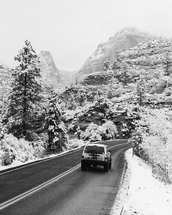 A white vehicle drives on a winding road through a snowy mountain landscape with pine trees.