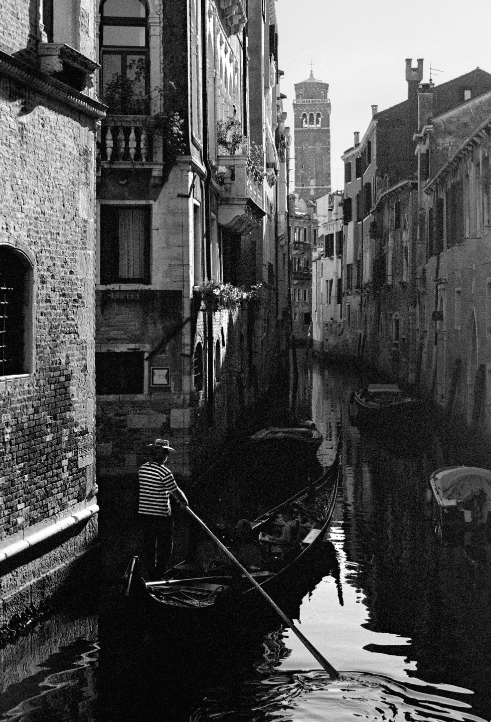 A gondolier stands in a gondola on a canal in Venice, with tall buildings and a bell tower visible in the background.