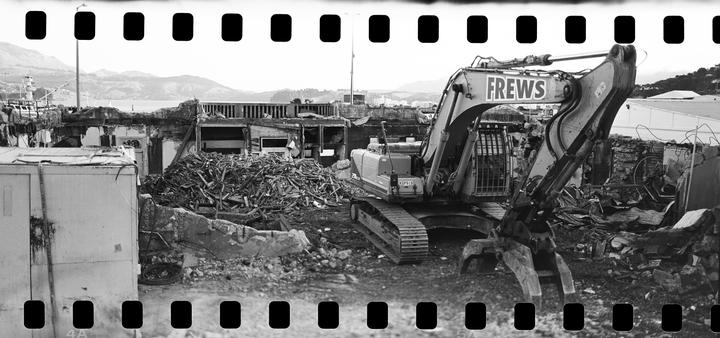 An excavator sits amidst the rubble of a demolished building with mountains and water in the background.