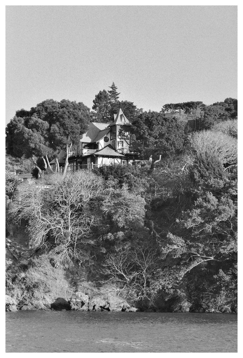 A large house with a turret is nestled among trees on a hillside overlooking water.